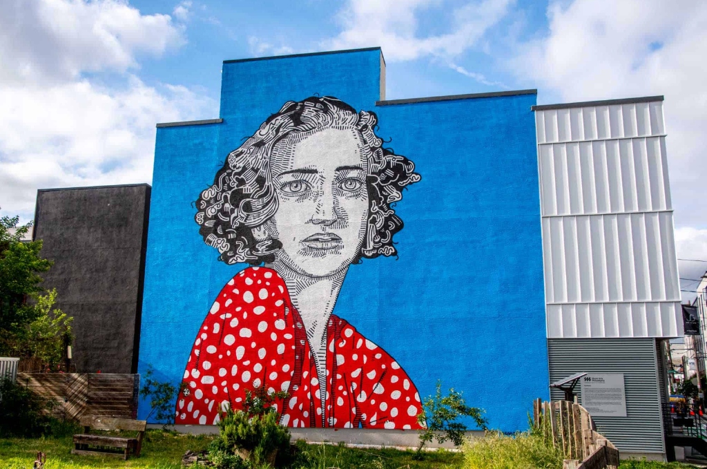 Persistence, a mural by Jason Andrew Turner: against a towering
                   bright blue wall, a figure with dark curled hair wears a red
                   dress patterned with white polka dots.